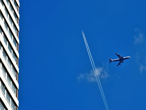 A jet plane with contrail flying high in the sky next to a modern skyscraper.