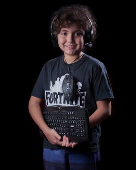 Young boy smiling while holding keyboard and wearing a gaming headset against dark background.
